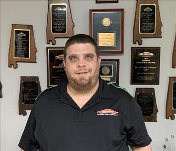 Photo of male employee standing in front wall with awards hanging and he is wearing a black collared shirt with SERVPRO logo